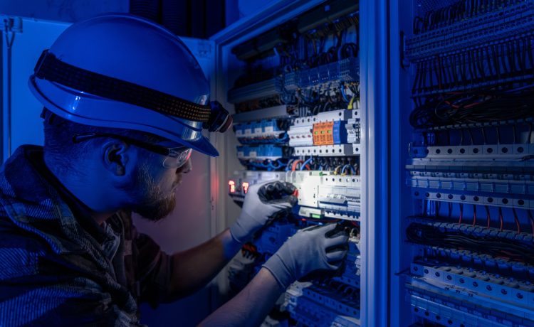 Male electrician working in switchboard with electrical connecting cable. Young adult electrical engineer in special clothes with flashlight on helmet in dark room with emergency lights in background.