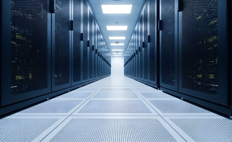 A long, symmetrical corridor stretches into the distance, flanked by tall, dark server racks with perforated fronts. The floor is tiled with a grid pattern of perforated squares. Overhead, rectangular fluorescent lights illuminate the space with a cool, white glow, creating a sterile and organized atmosphere. The perspective draws the viewer down the aisle towards a bright, white opening at the far end.